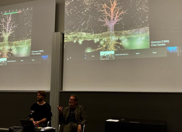 Two people, Jakob Kudsk and Mogens Jacobsen, are sat on chairs in a lecture room. Behind them is a projection of the Zoom call where one of Char Davies' art works is displayed. 