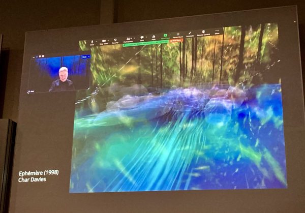 A Zoom call with Char Davies in a little square in the corner and one of her virtual art pieces showing on the screen in beautiful fluorescent greens and blues layering on top of forrest. 